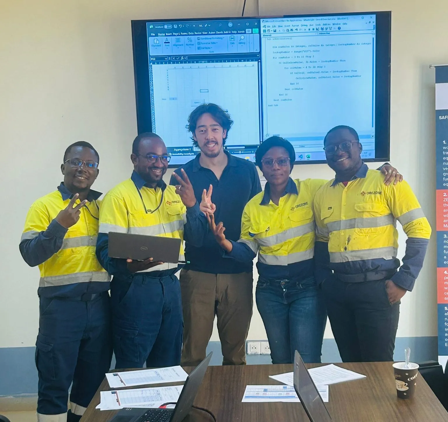 Group of five people in high-visibility yellow workwear posing together in front of a screen displaying engineering or programming software, with one person holding a laptop and others making peace signs, in a training setting.
